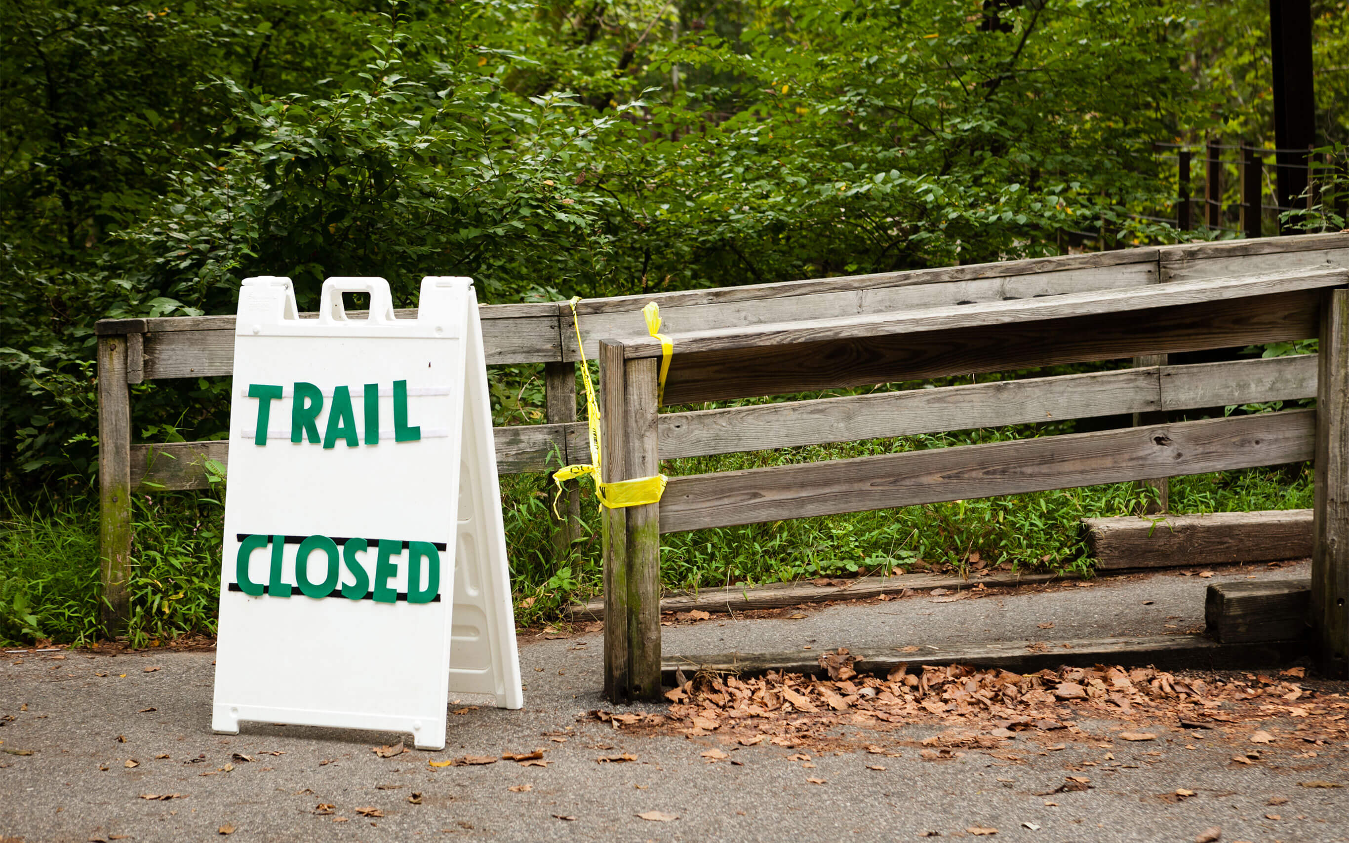 An outdoor path blocked with caution tape and a sign reading Trail Closed